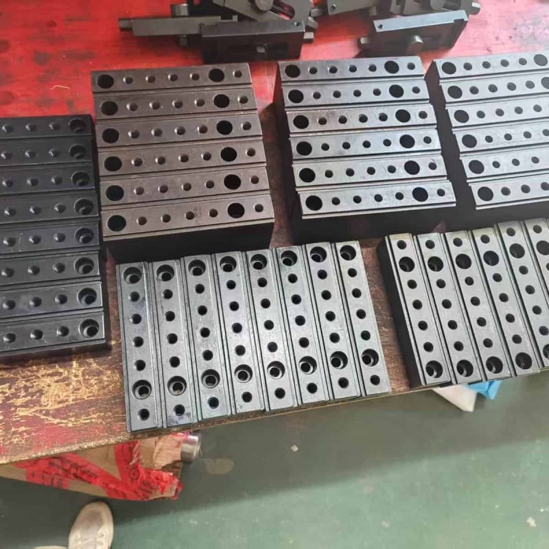 Precise black metal mechanical components with multiple drilled holes, arranged on a wooden workbench for CNC spring manufacturing processes.